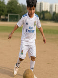 Child in a soccer uniform kicking a ball on a field with buildings in the background